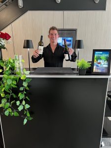 A smiling bartender holding bottles of wine at a mobile bar setup in an office.